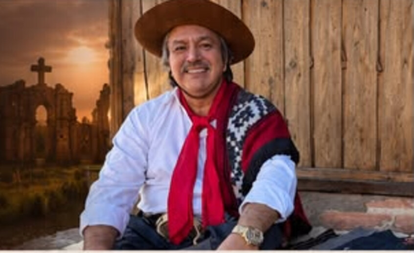 Jorge Guedes sentado em frente a uma parede de madeira, com as ruínas de São Miguel das Missões ao fundo no pôr do sol