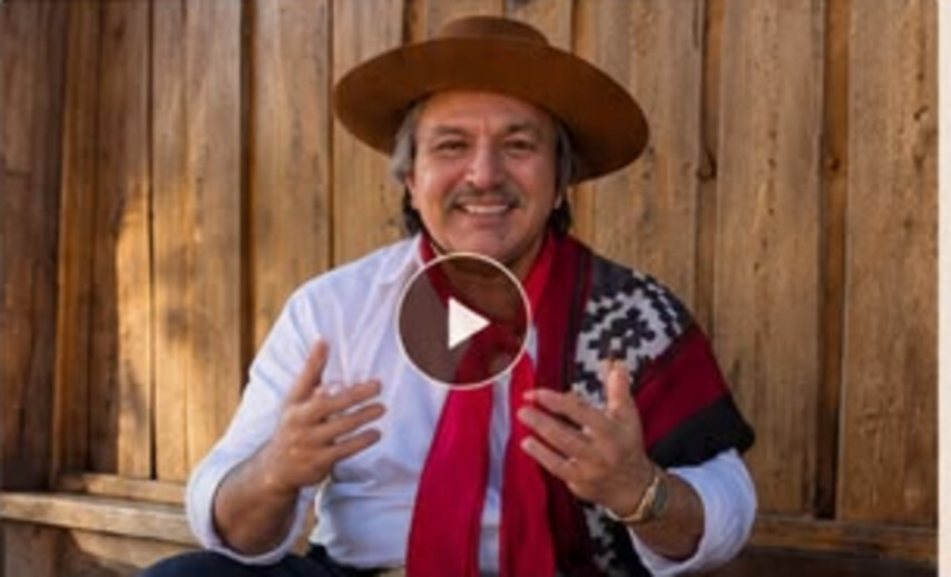 Jorge Guedes sorrindo, de chapéu e lenço vermelho, em frente a uma parede de madeira